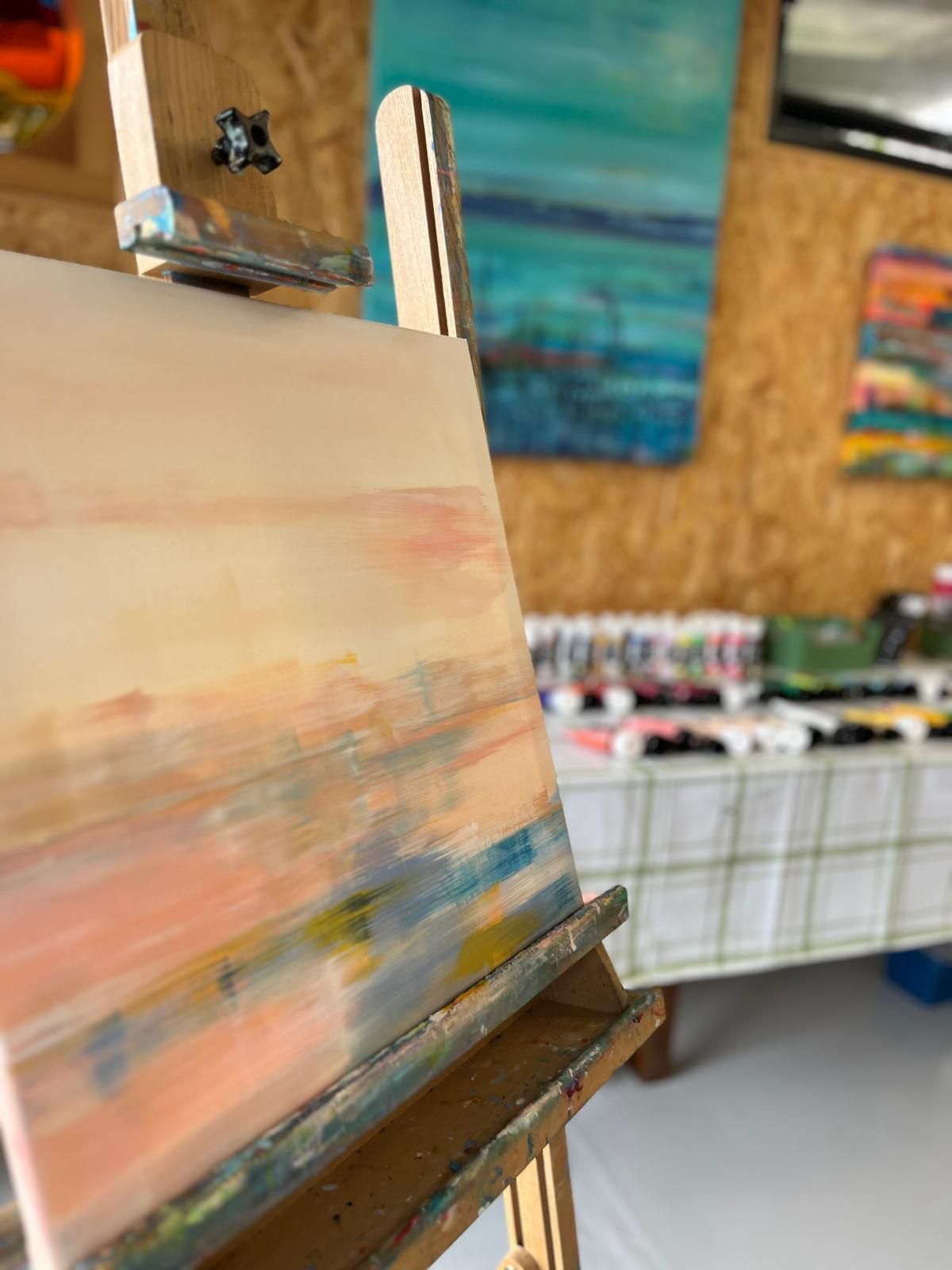 Painting on an easel with abstract strokes of peach, blue, and yellow. Other paintings and art supplies in background.