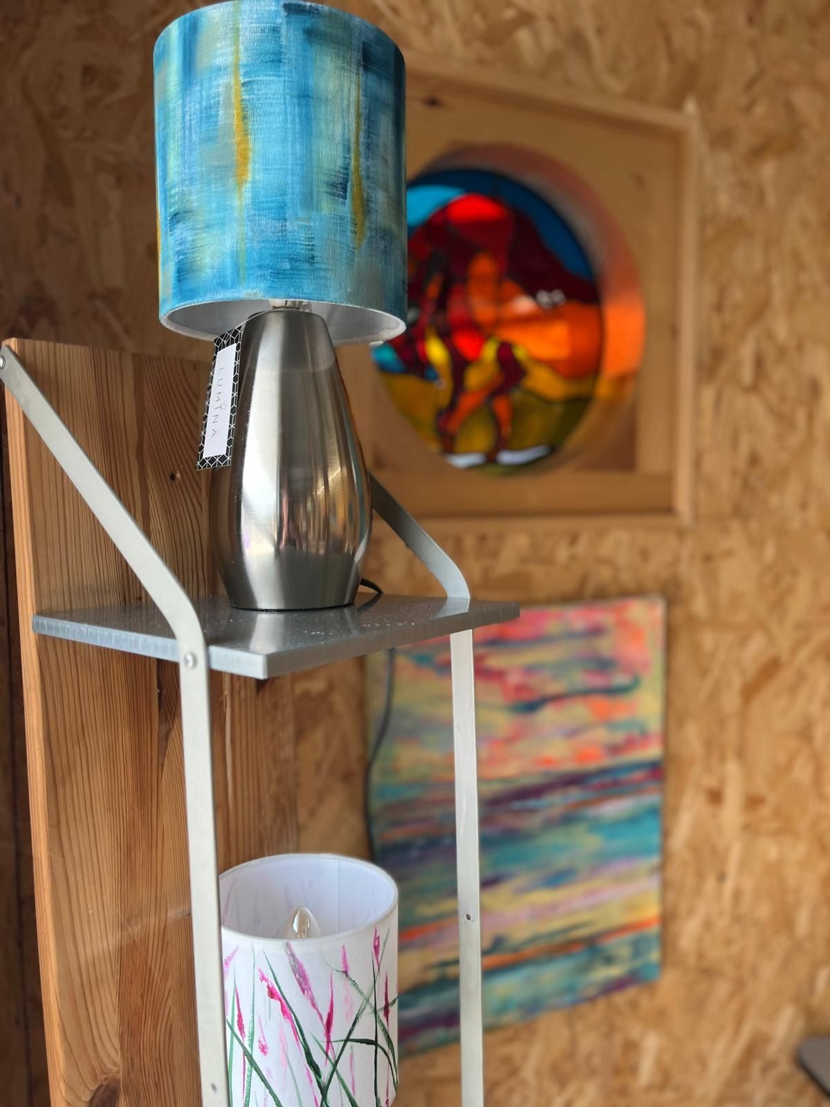 A lamp with a blue lampshade sits on a shelf. Artwork and a second lamp are visible.