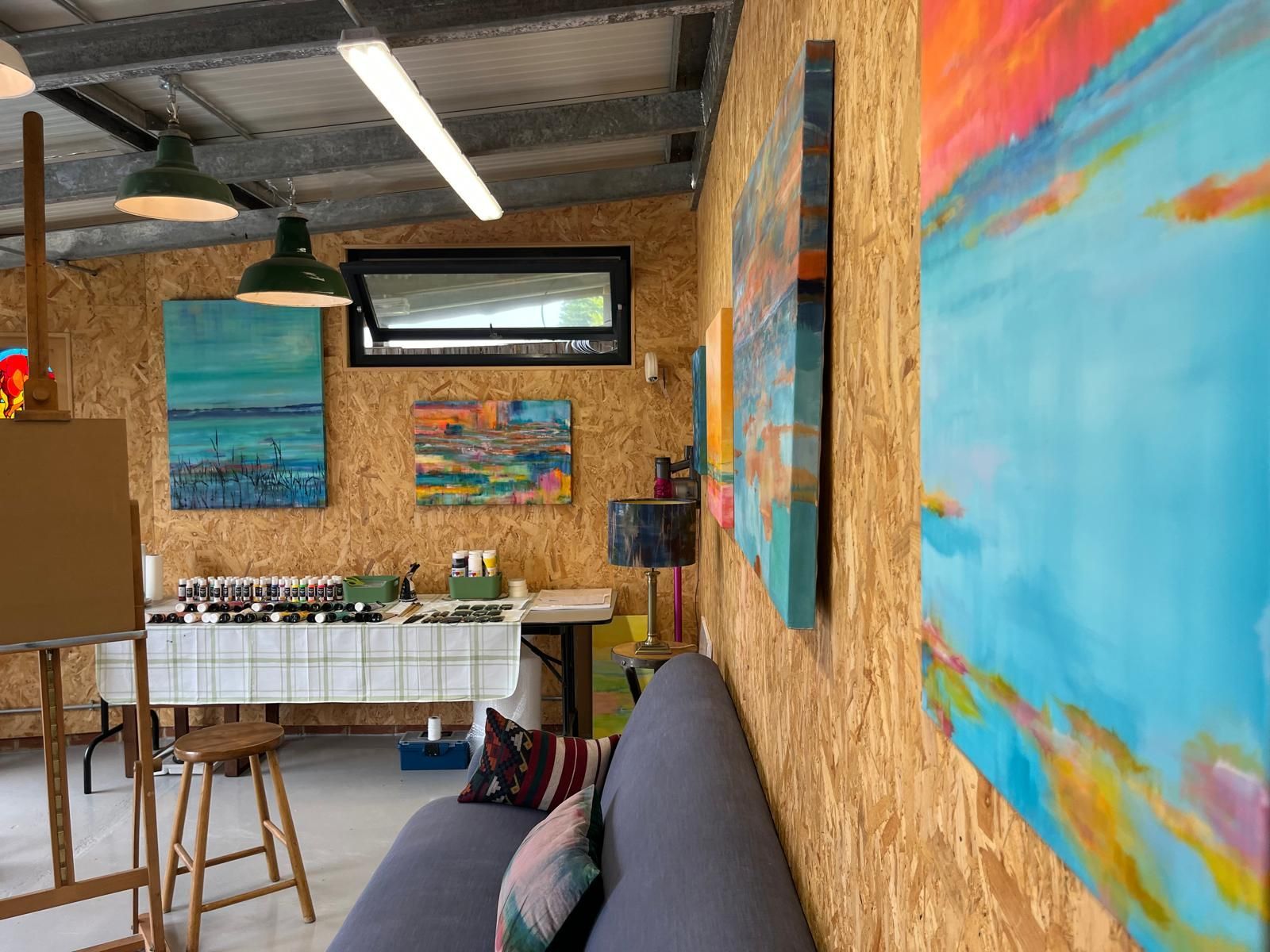 Art studio interior with paintings on walls, easel, paint supplies, and a couch.