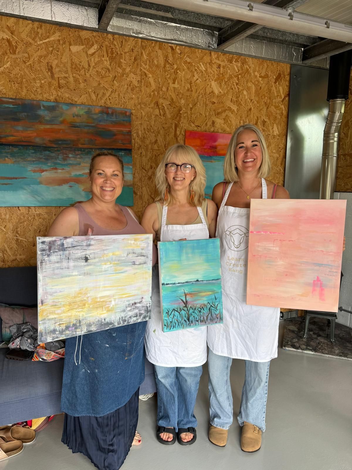 Three women in paint aprons hold up abstract paintings in an art studio.