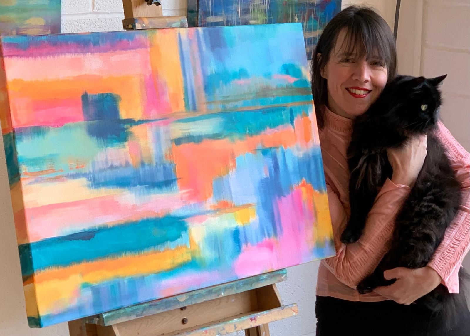 Jemsy holding a black cat next to colorful abstract painting on an easel.