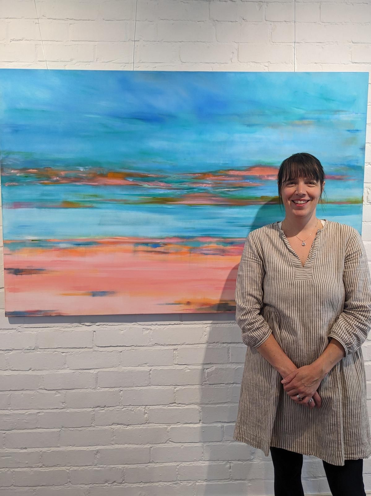 Jemma stands next to a large abstract painting of blue, orange, and pink hues. White brick wall.
