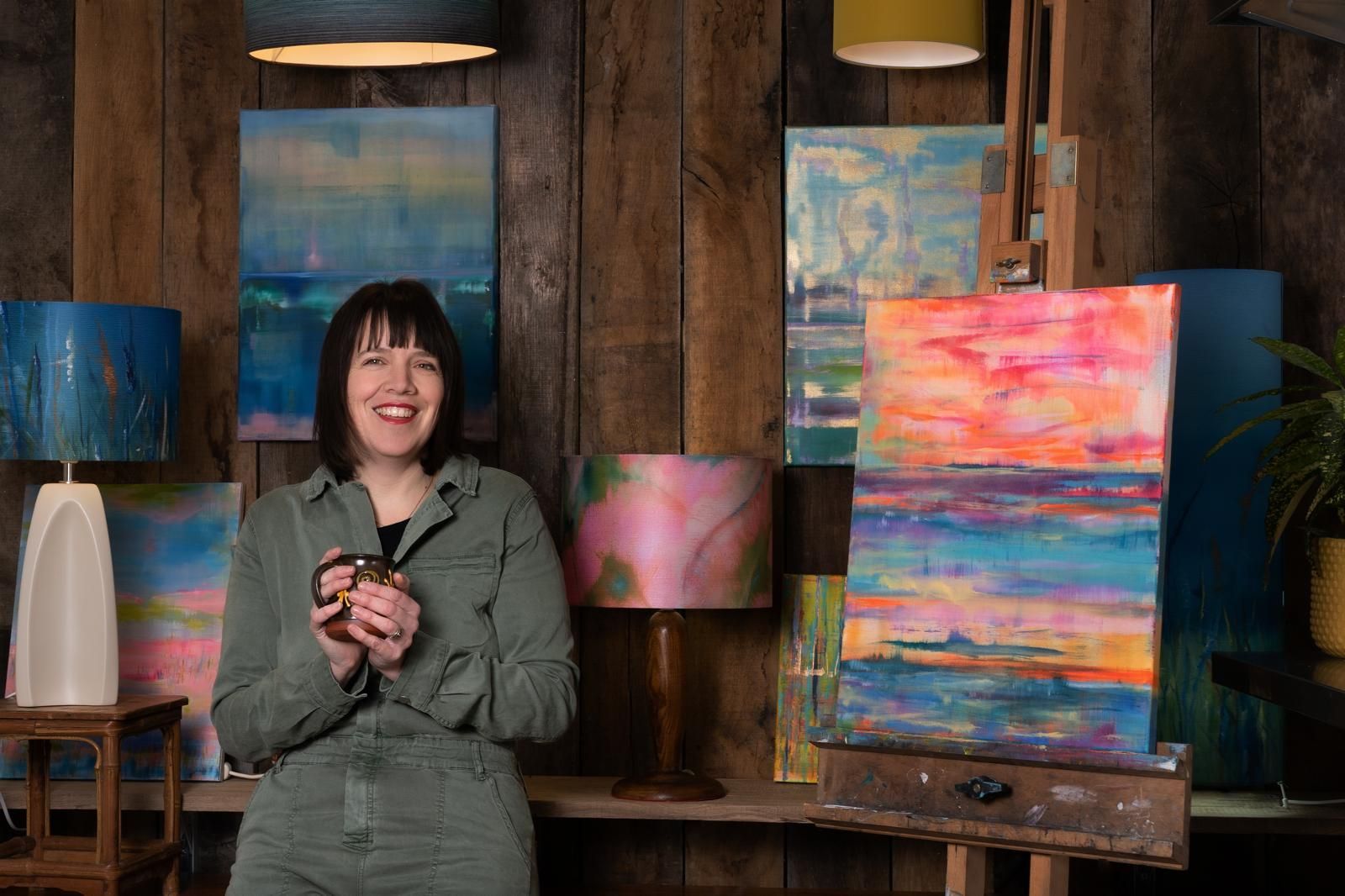 Jemma Jones an artist in studio smiles, holding a mug, surrounded by colorful abstract paintings.