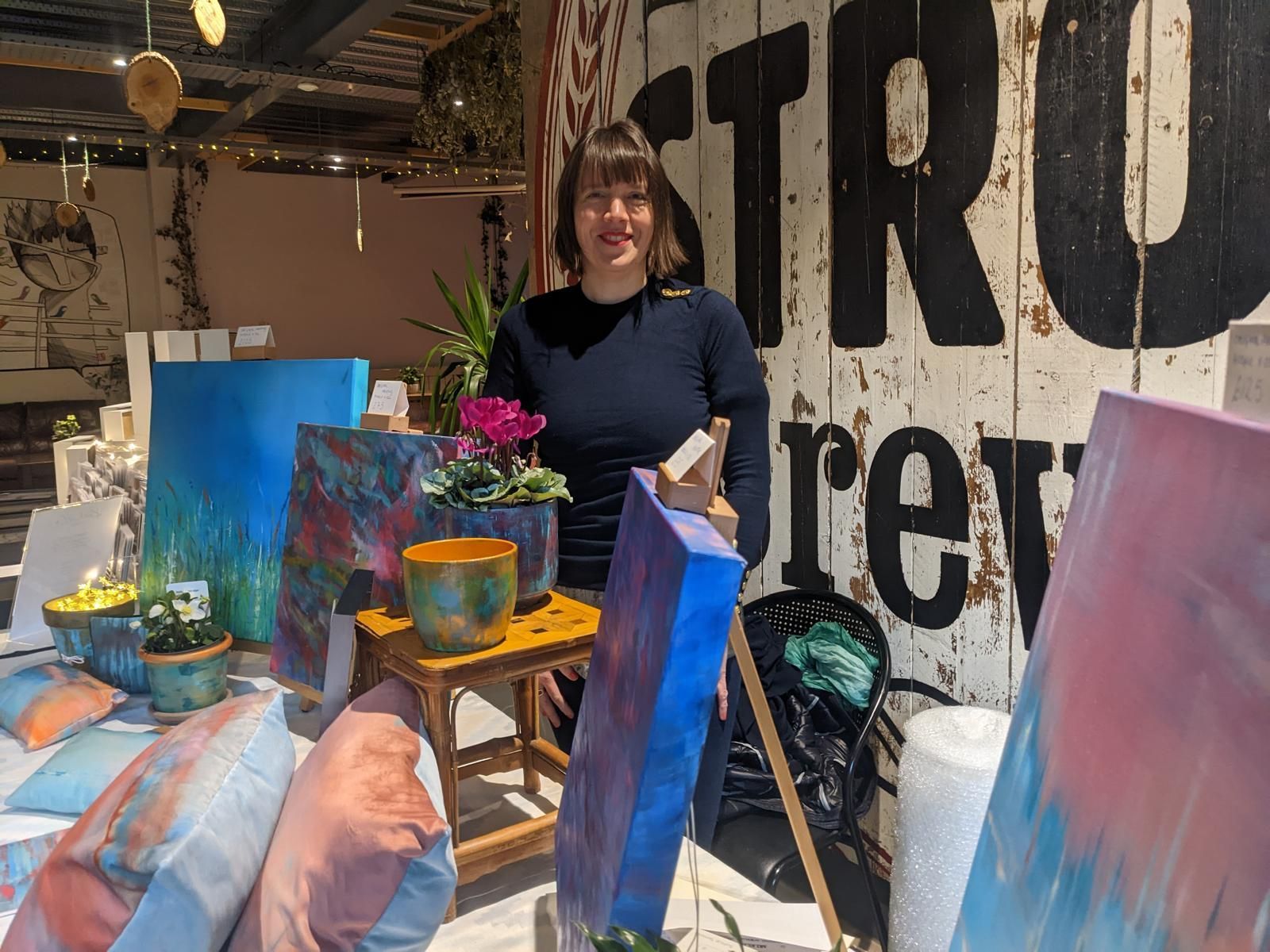 Jemma at art market booth, surrounded by colorful paintings and pottery.