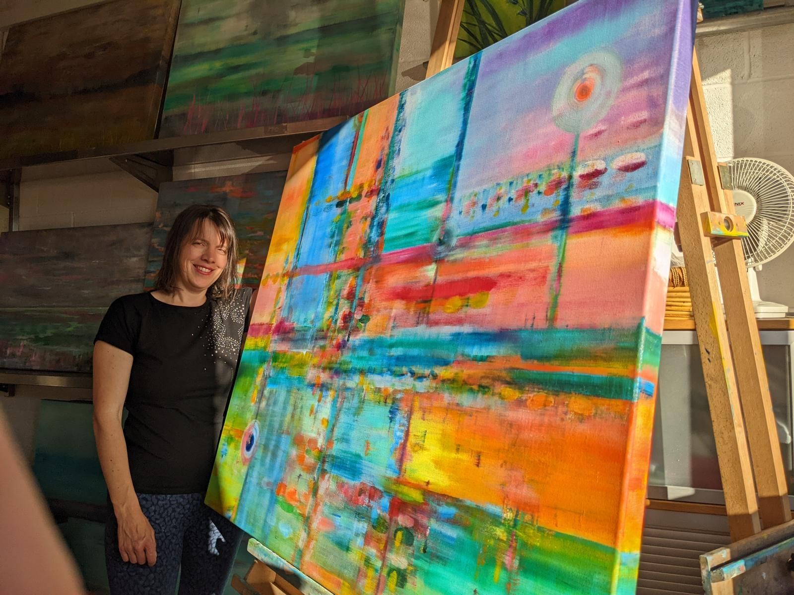 Woman in black shirt smiles, holding colorful abstract painting in art studio.