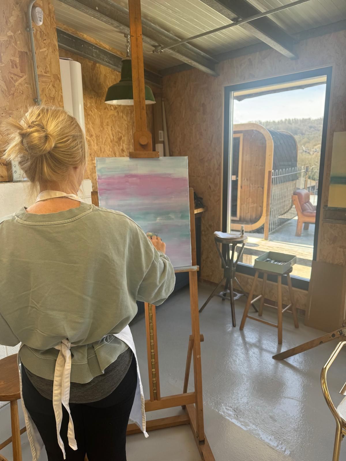 A workshop customer painting abstract art in a studio. Light pink and blue colors on canvas. Studio has outdoor view.