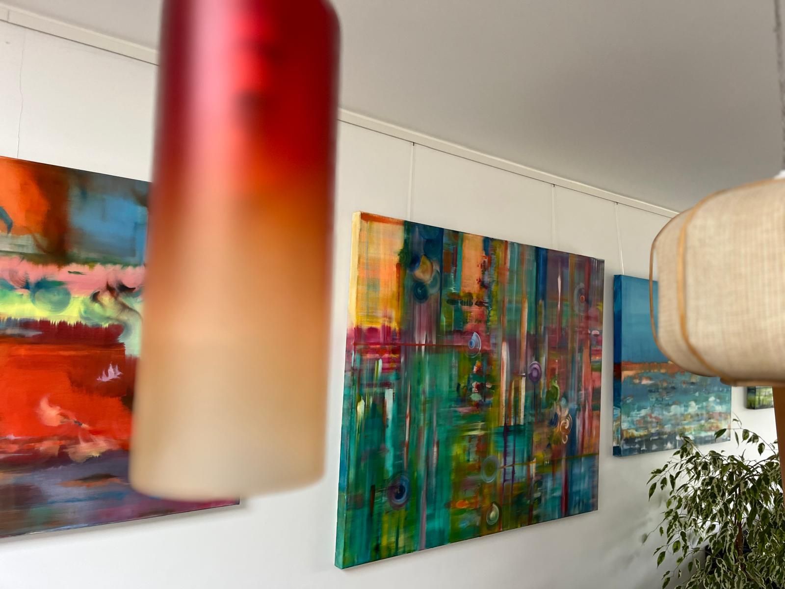 Abstract art on white wall, colorful paintings lit by gradient red and tan lamp, and light fixture.