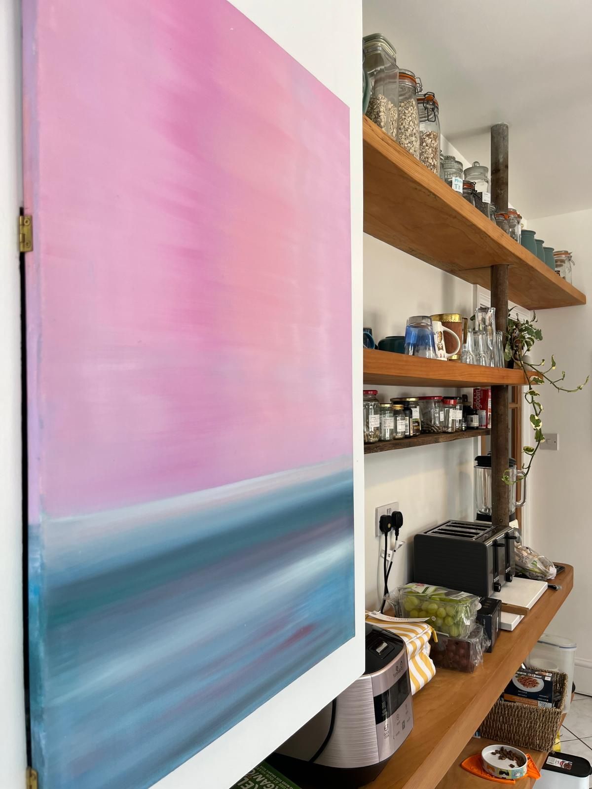 Pink and teal abstract painting on a white wall, next to wooden shelves holding jars and kitchen items.