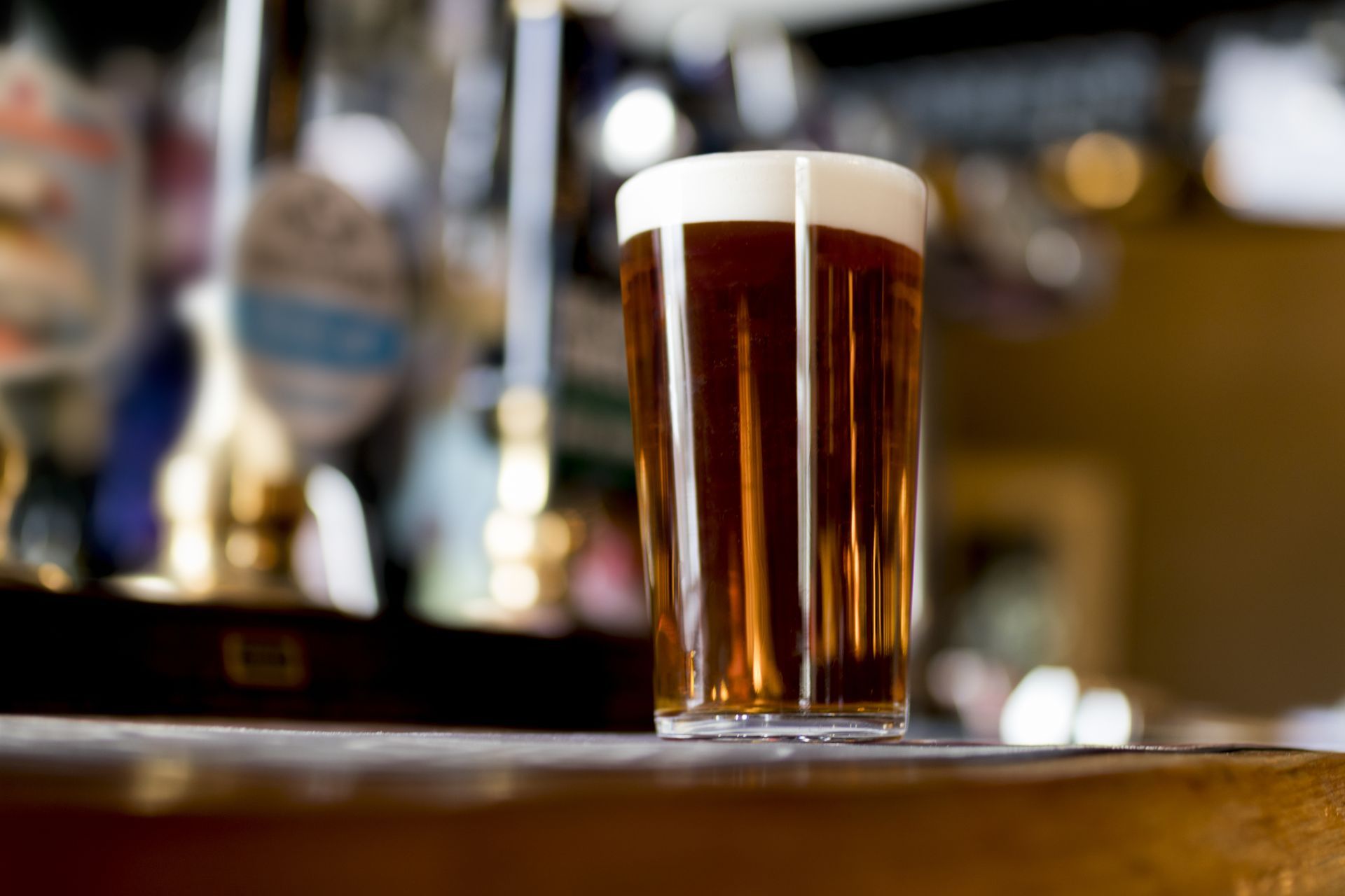 A pint of dark beer with a white head on a wooden bar, pub background.