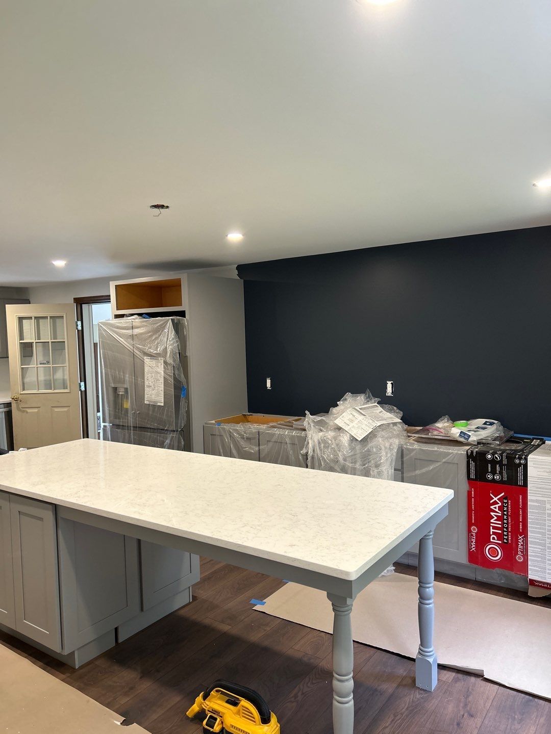 Kitchen under renovation. Gray cabinets, white countertop island, dark blue wall, stainless fridge, and various construction materials.