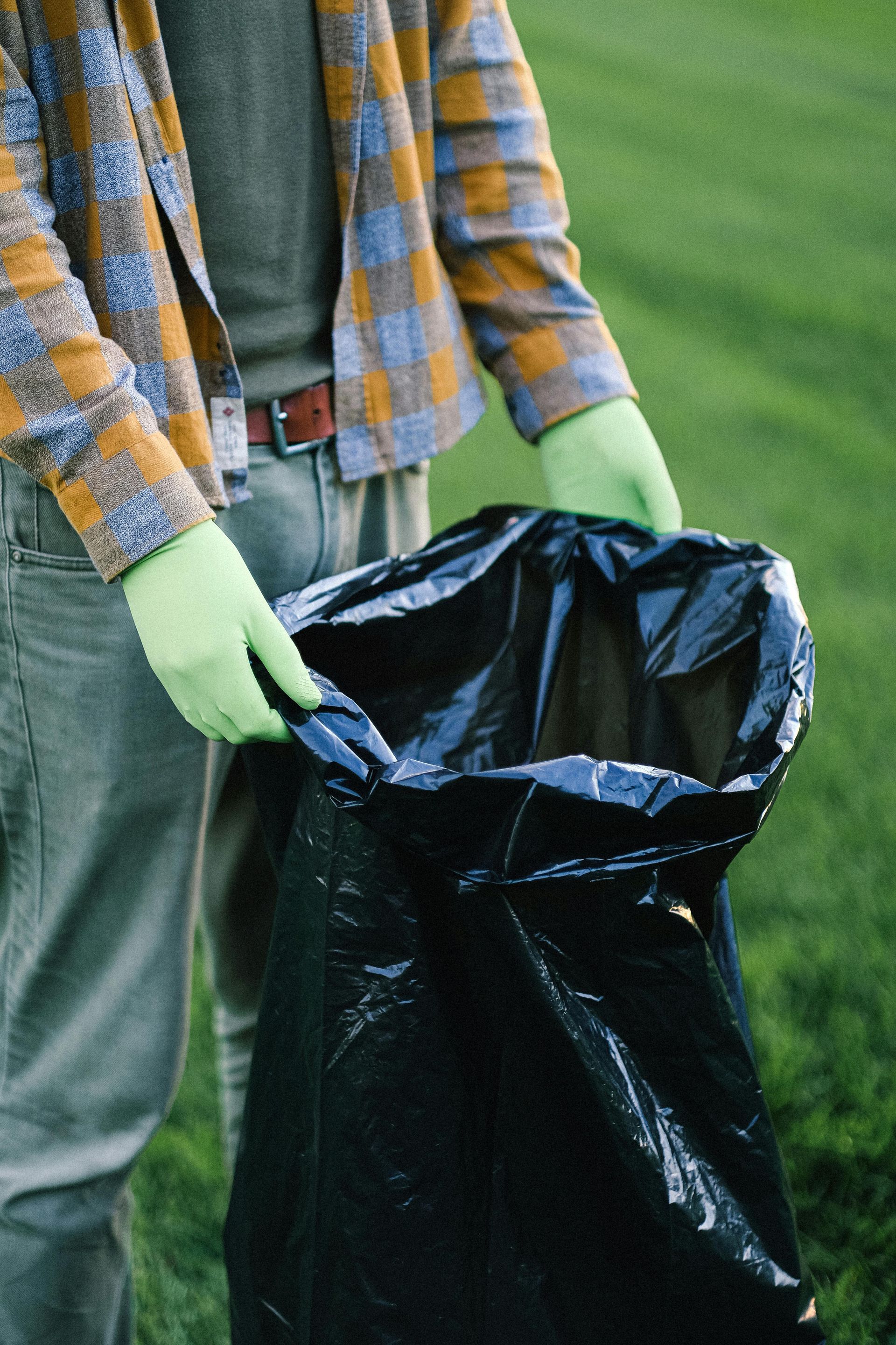 Person holding open black trash bag outdoors, wearing plaid shirt and gloves.