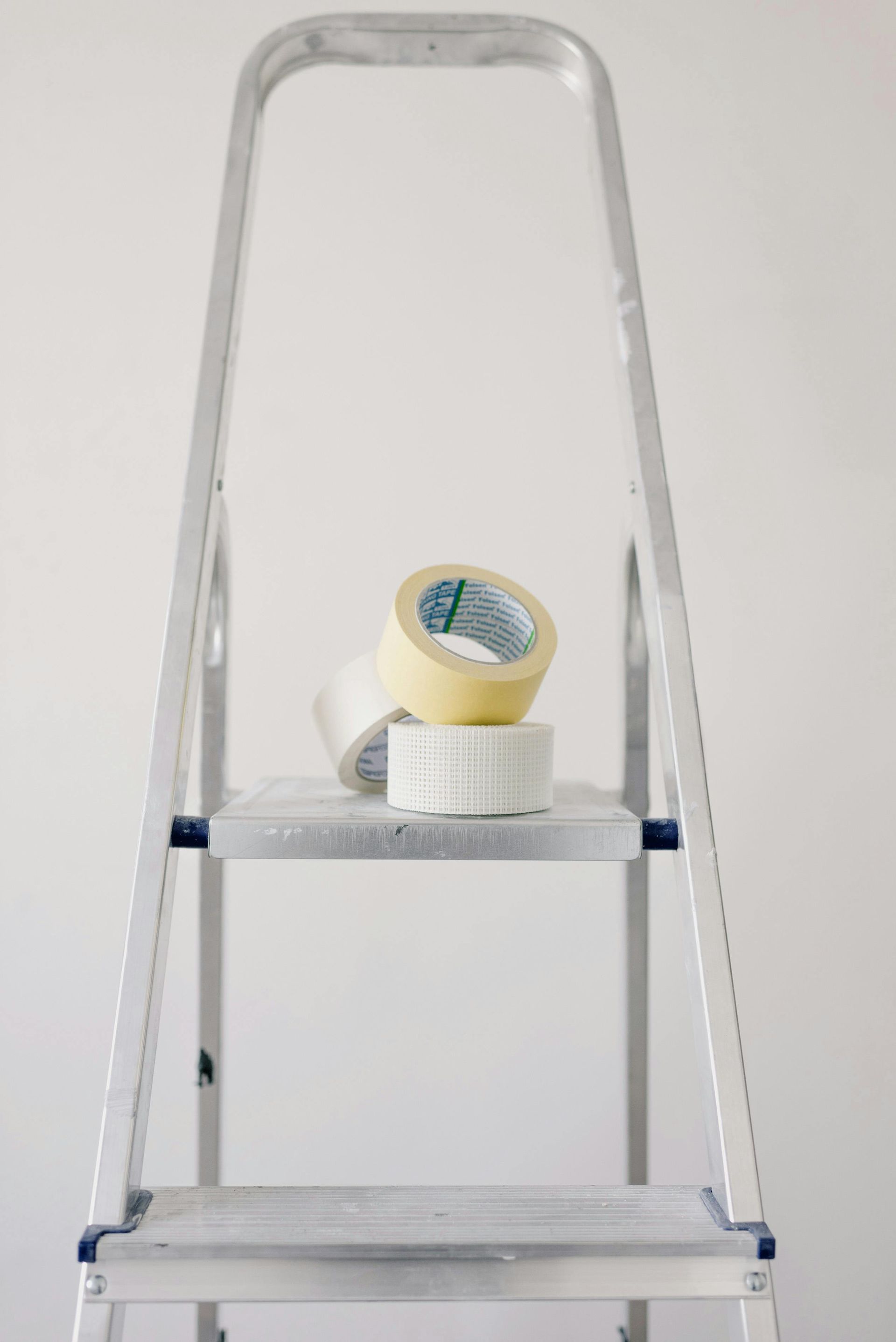 Aluminum step ladder with three rolls of tape on a shelf, against a white wall.
