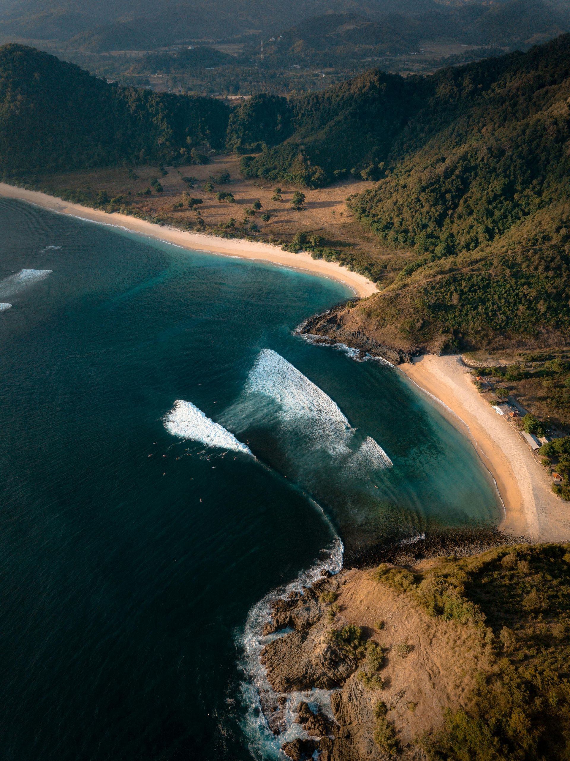 An aerial view of a beach with mountains in the background and waves crashing on the shore.