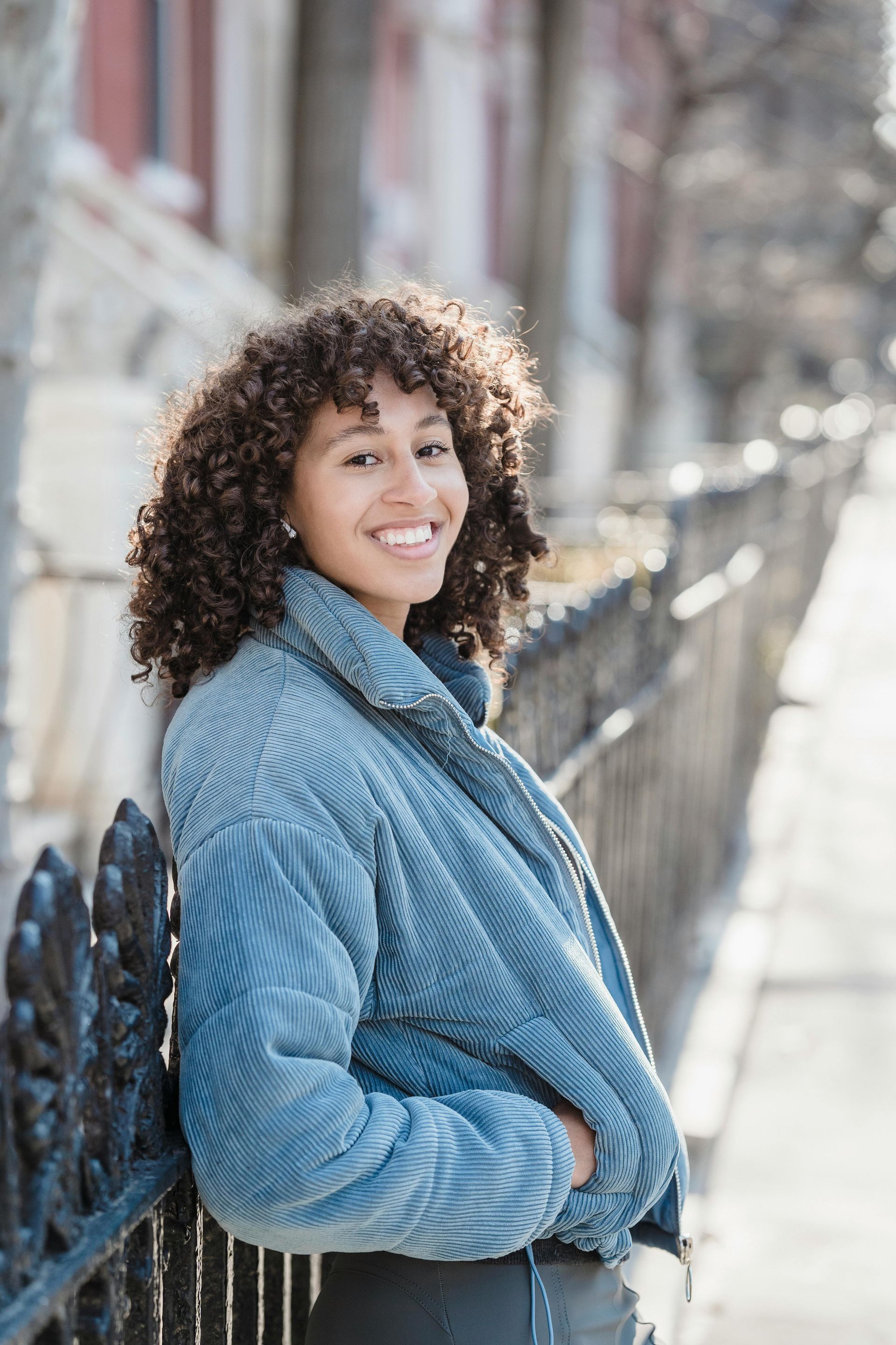 A woman with curly hair is leaning against a fence and smiling.