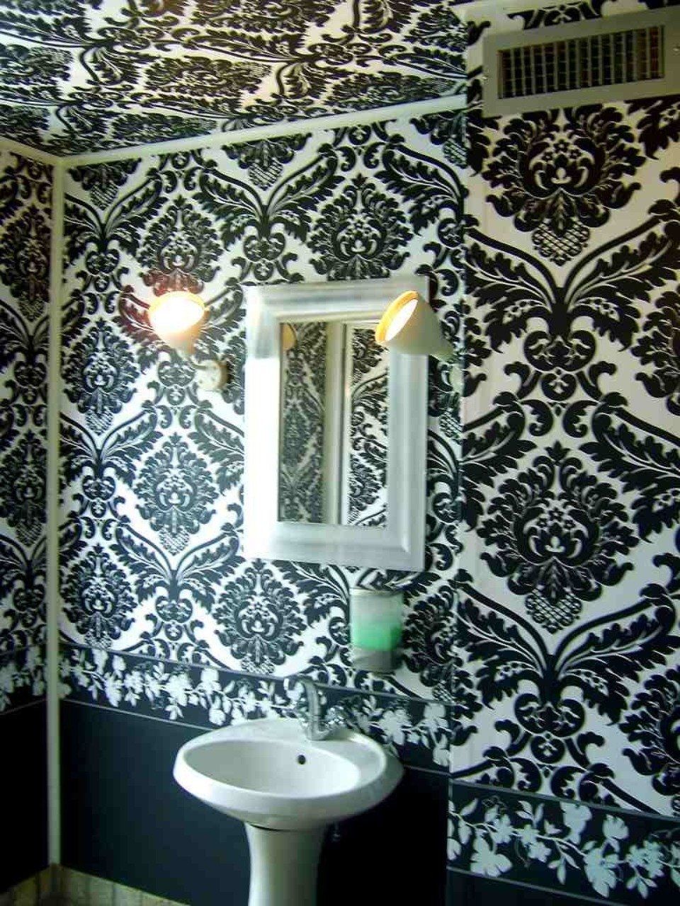 Bagno con carta da parati damascata bianca e nera, lavandino bianco, specchio e due luci.