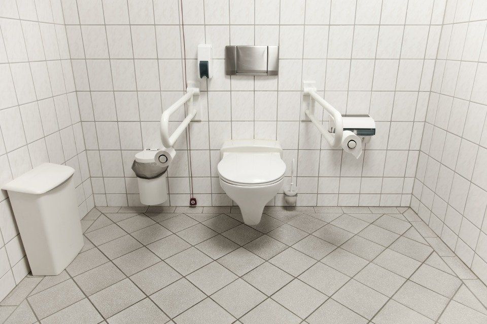 Bagno accessibile con WC, maniglioni e cestino per i rifiuti. Pareti e pavimento sono rivestiti di piastrelle bianche.