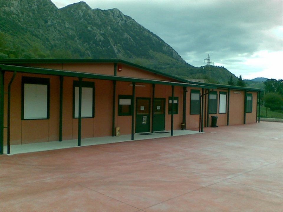 Un edificio color marrone chiaro, a un solo piano, con le montagne sullo sfondo e un cielo nuvoloso.