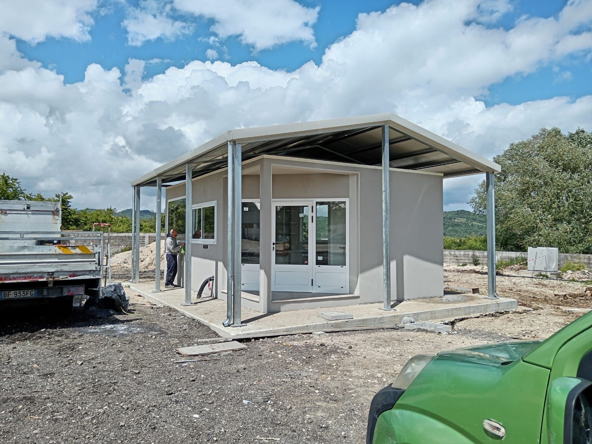 Edificio grigio con tetto e supporti in metallo. Porte e finestre aperte. Cielo nuvoloso. Veicolo verde in primo piano.