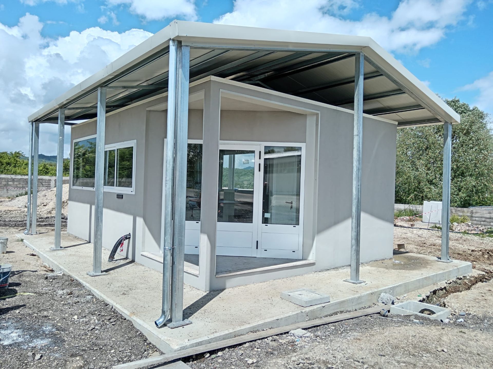 Edificio grigio con tenda da sole in metallo, finestre e porte, su una lastra di cemento in un ambiente esterno.