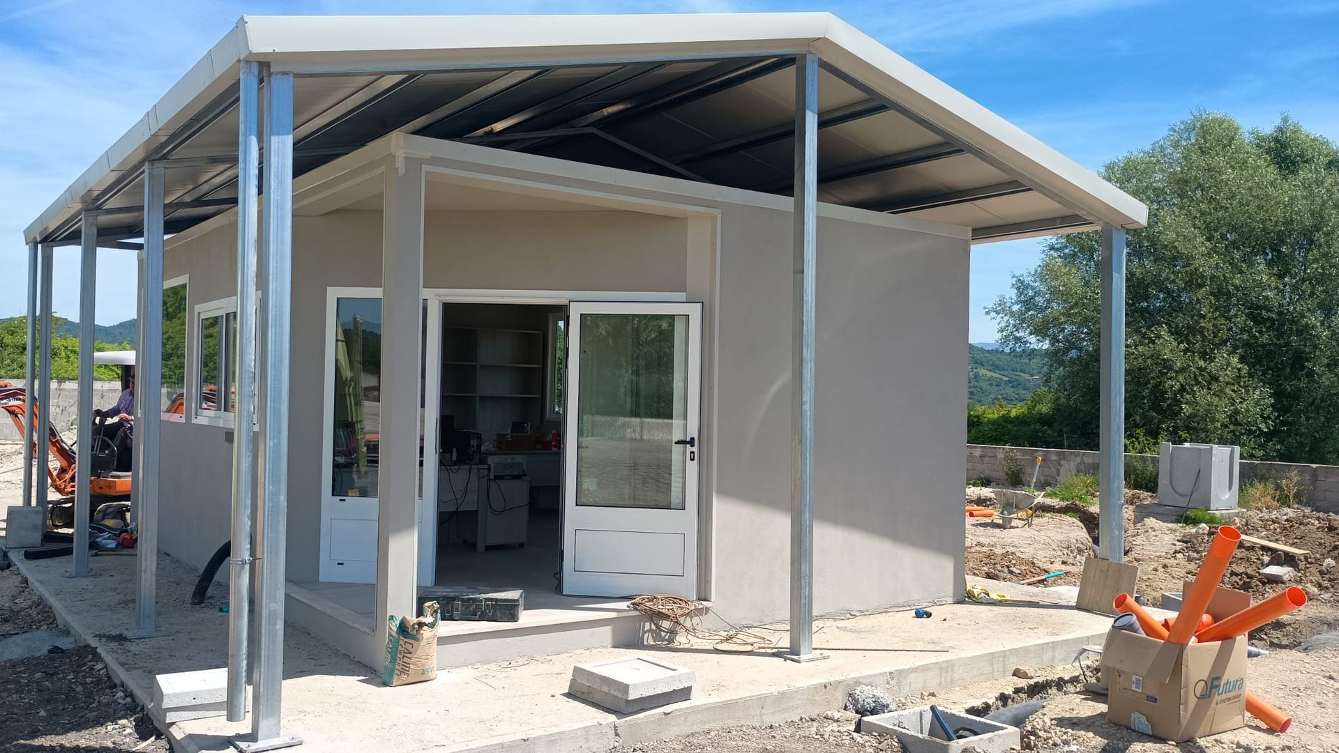 Piccolo edificio grigio chiaro con tetto e portico in acciaio; in costruzione, immerso nella natura.