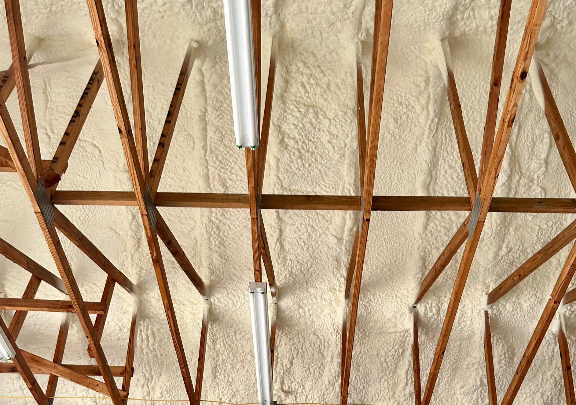 Attic rafters with spray foam insulation, beige with wooden beams. Two fluorescent lights hang down.