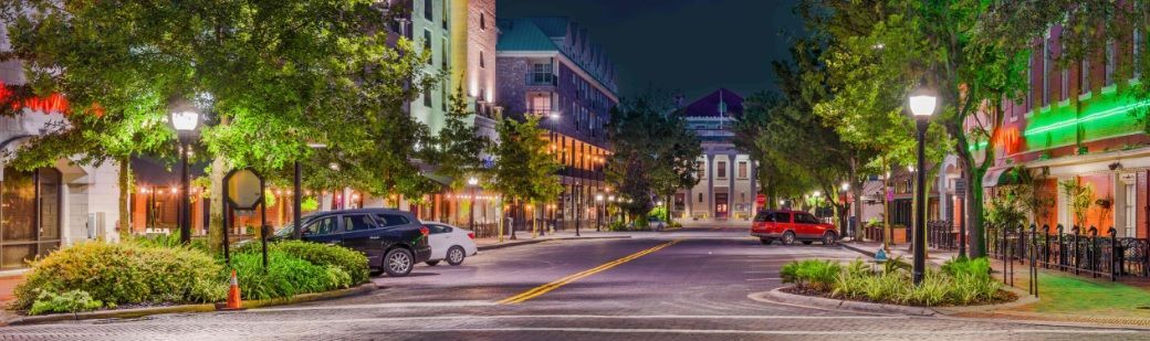 Night street scene with lit shops, trees, and streetlights. A car is visible in the middle.