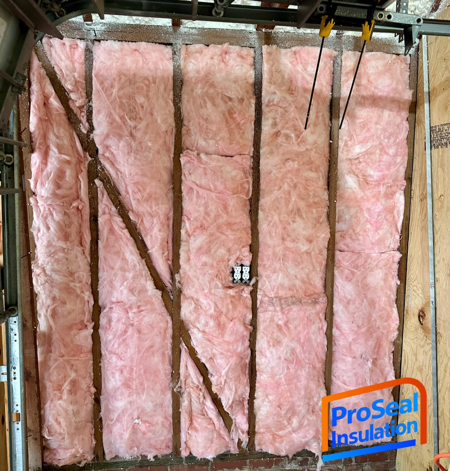 Pink fiberglass insulation installed between wooden studs in a wall, with an electrical outlet visible.