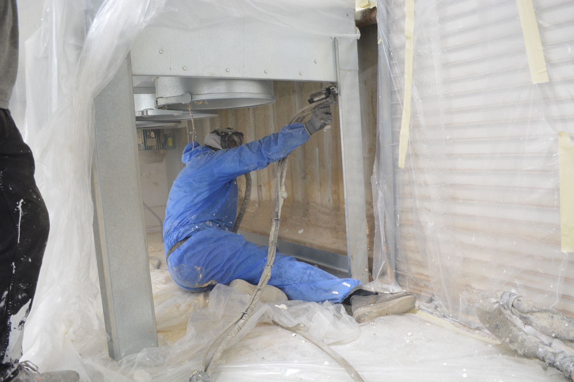 A person in a blue jumpsuit sprays paint inside an enclosed area. Protective plastic covers the surrounding space.