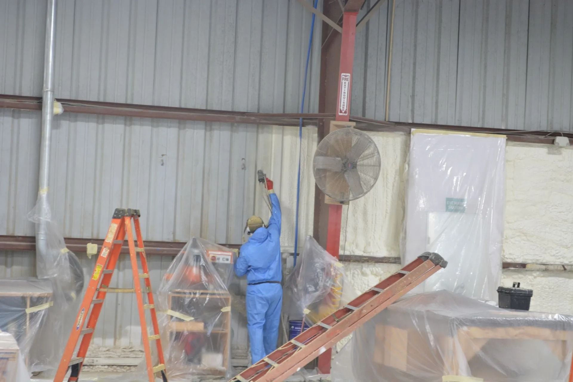 Person in blue jumpsuit sprays paint inside a warehouse, near a ladder and fan.