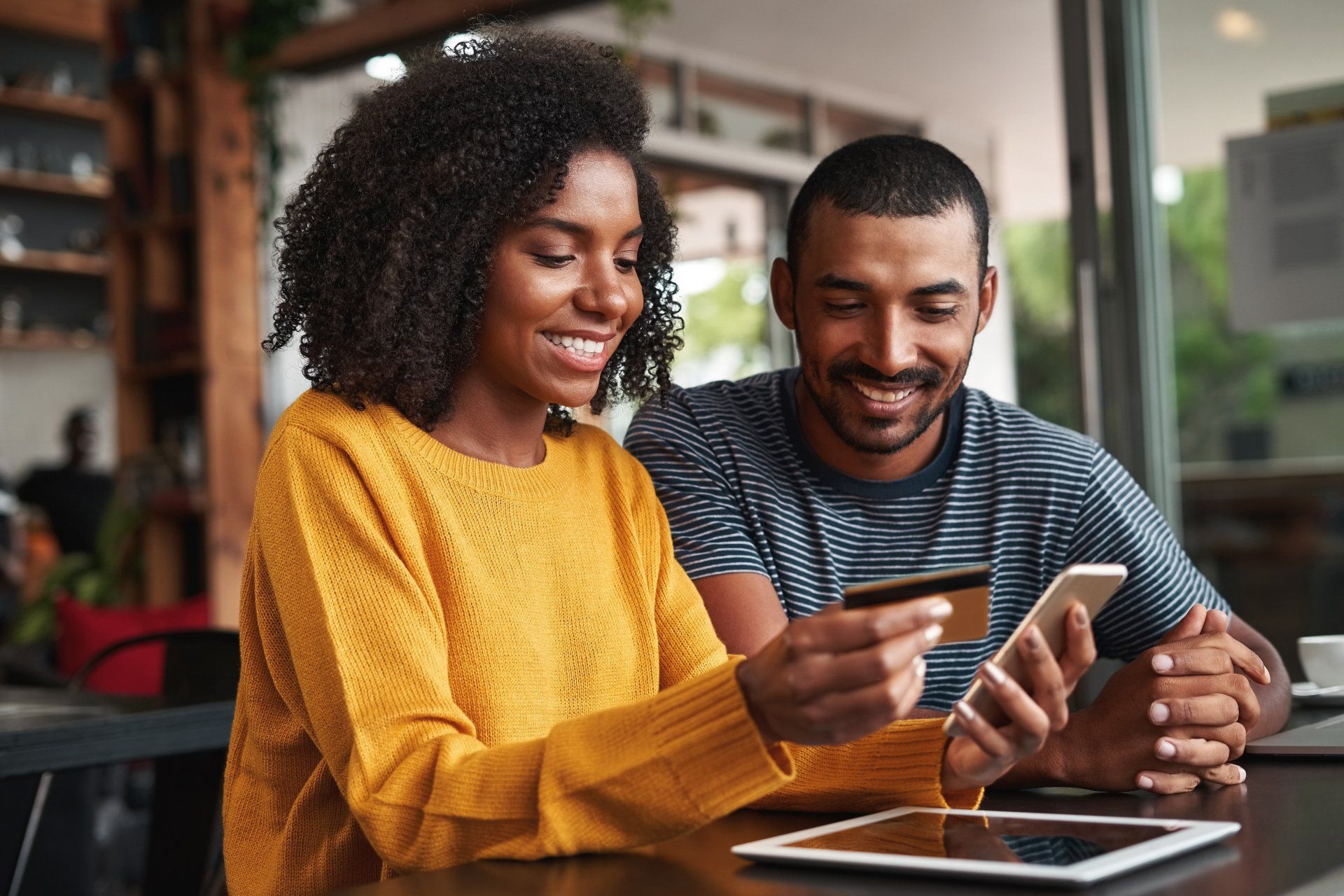 A man and a woman are sitting at a table looking at a tablet and a cell phone.