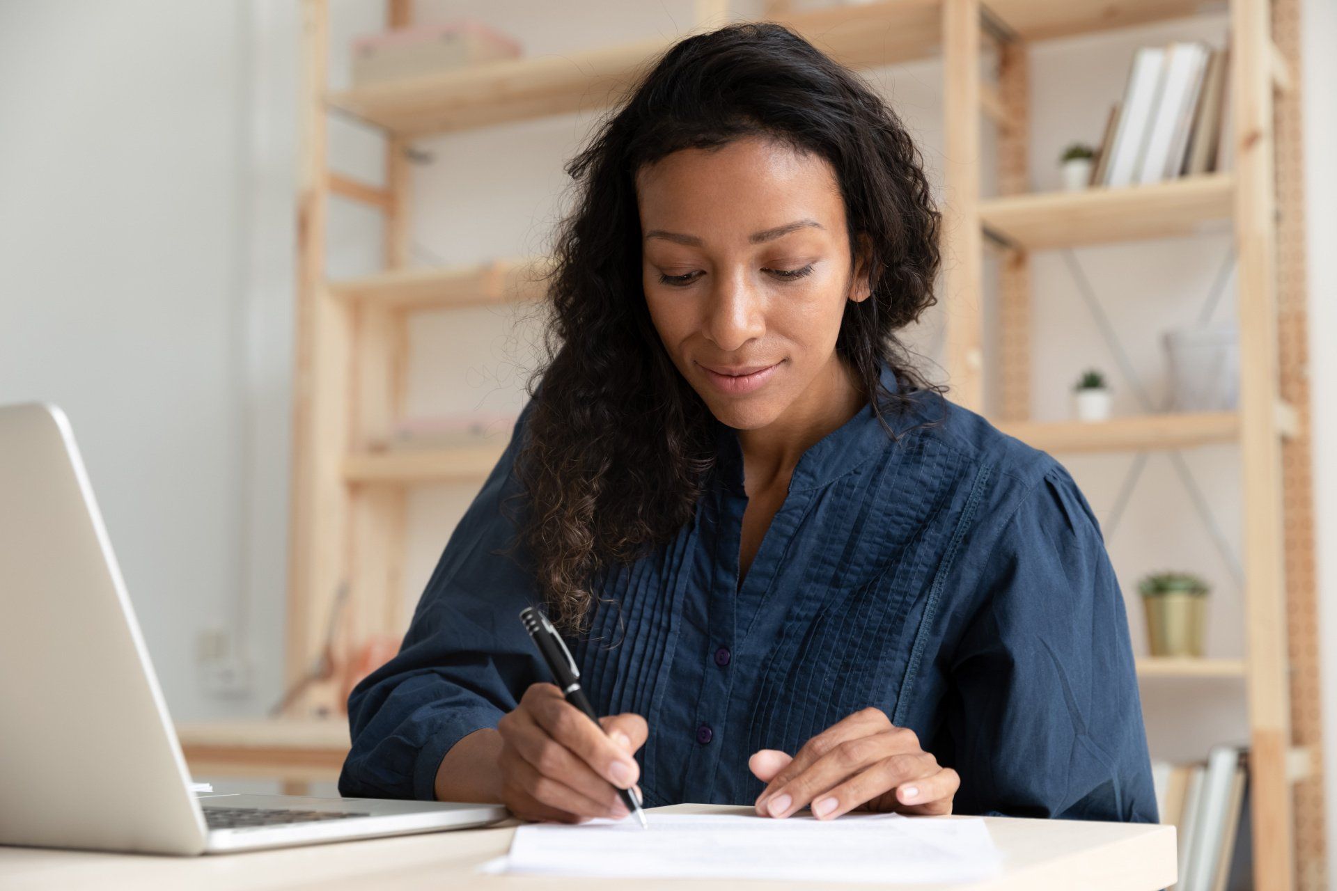 A woman is sitting at a desk with a laptop and writing on a piece of paper.
