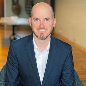 A bald man in a suit and white shirt is sitting in a chair.