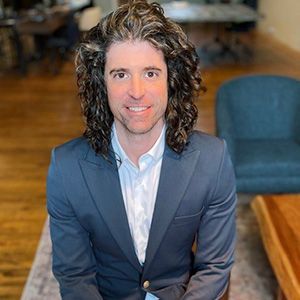 A man with long curly hair is wearing a suit and smiling for the camera.