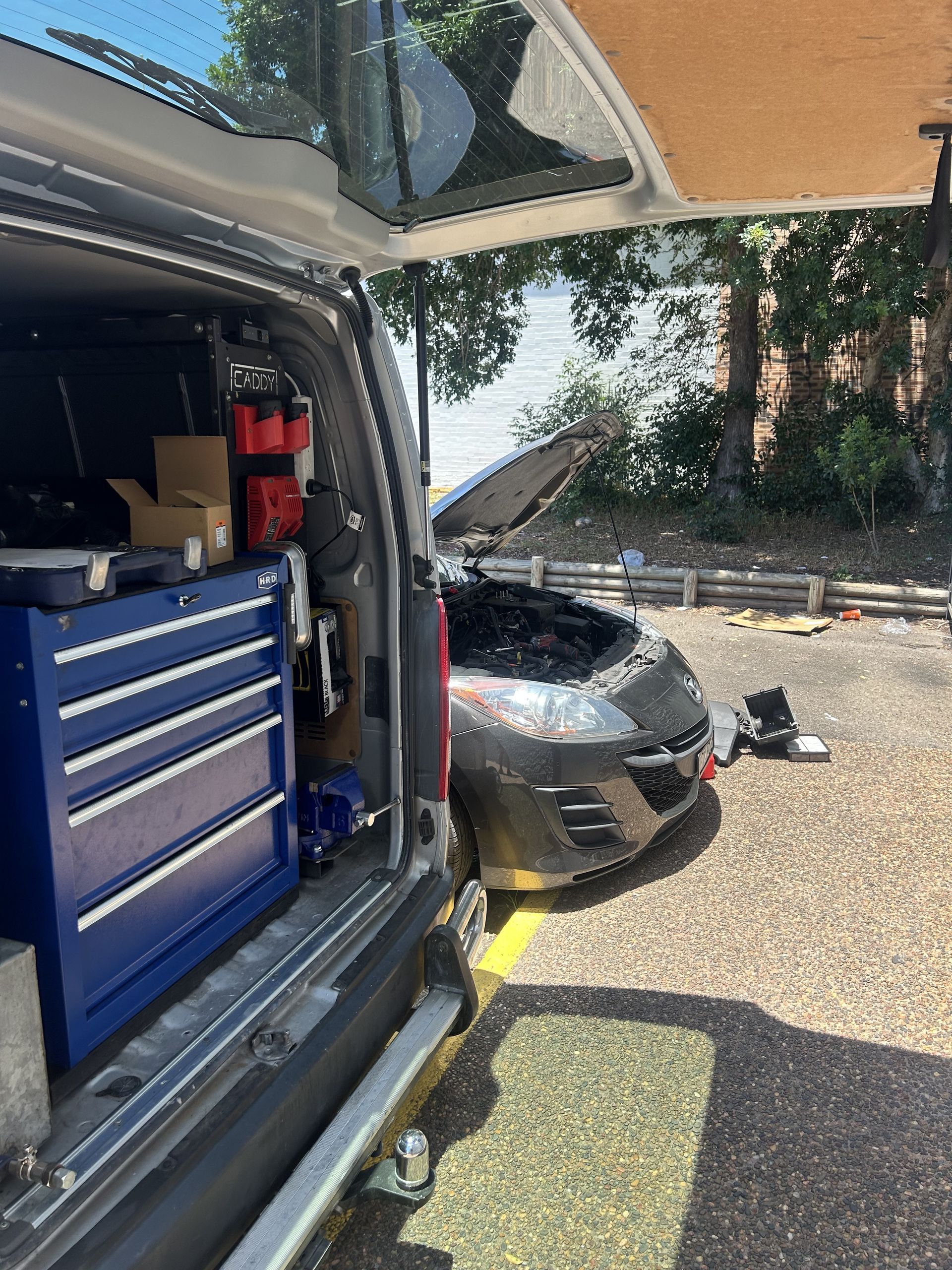 A mechanic's van open next to a car with an open hood, parked in a lot. Tools visible. — All Auto & Mechanical in Erina, NSW