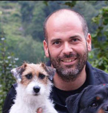 Un homme souriant tient un petit chien blanc et marron, ainsi qu'une partie d'un chien noir. En arrière-plan : des arbres.