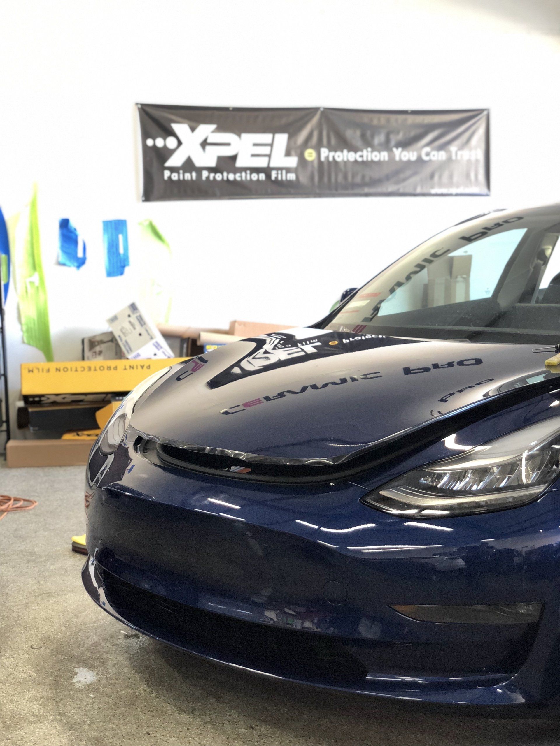 A blue tesla model 3 is parked in front of a xpel banner