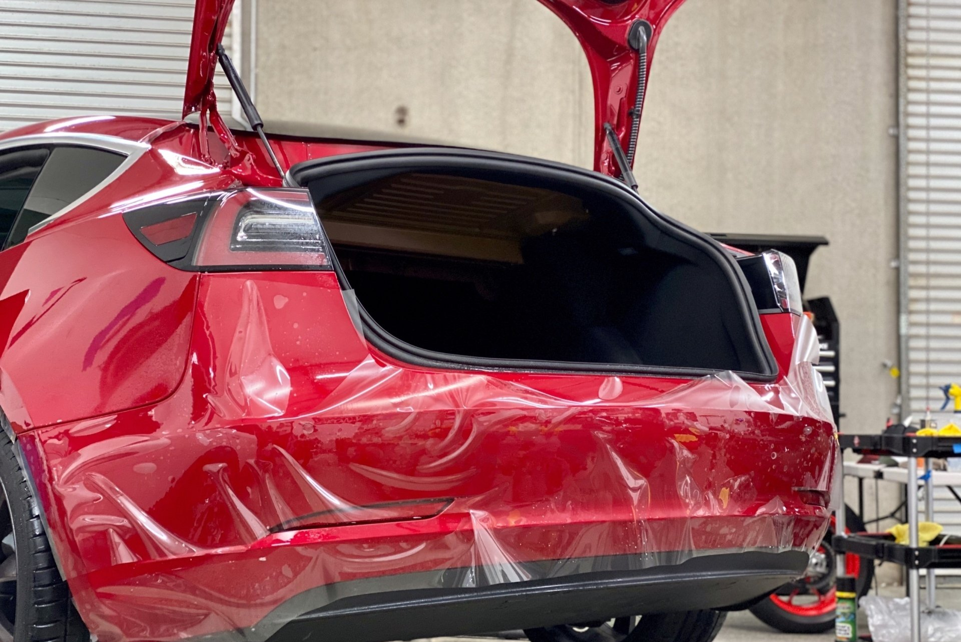 A red tesla model 3 with its trunk open is wrapped in plastic.