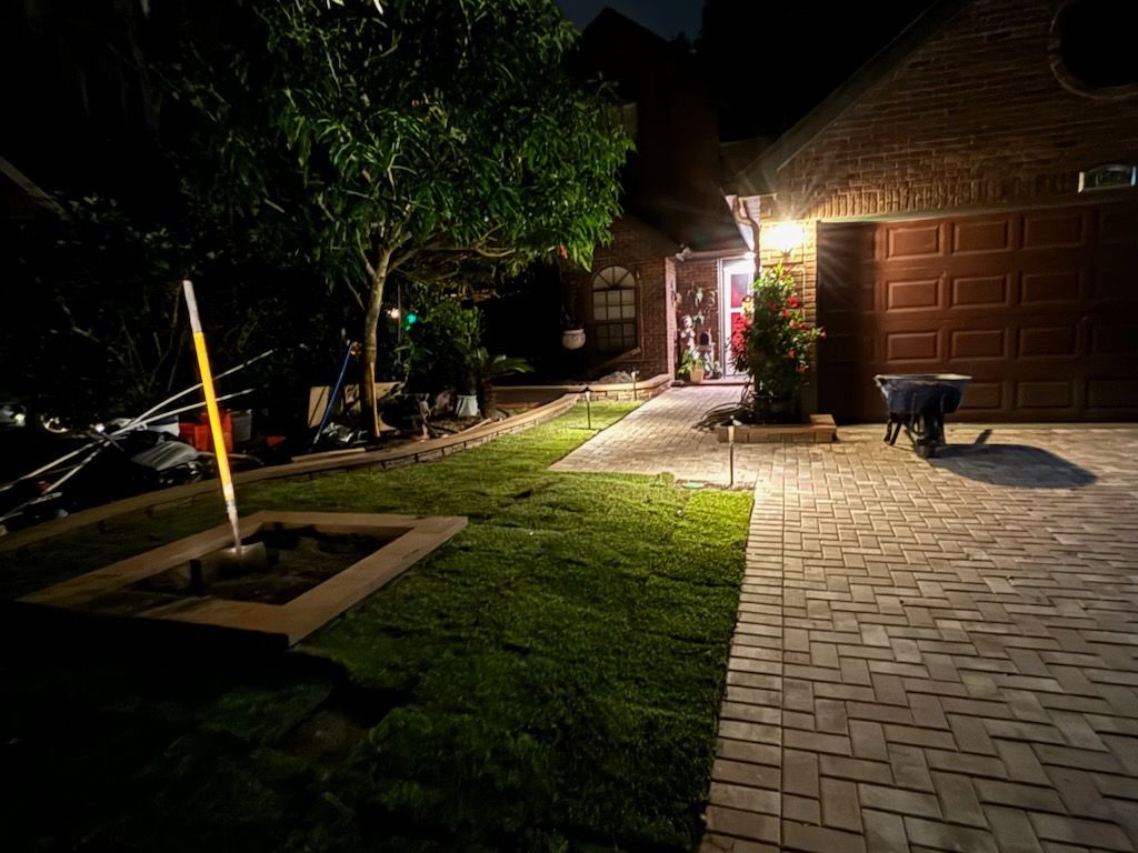 Night view of a house with a lit pathway, fresh lawn, and garage. A person stands in the doorway.