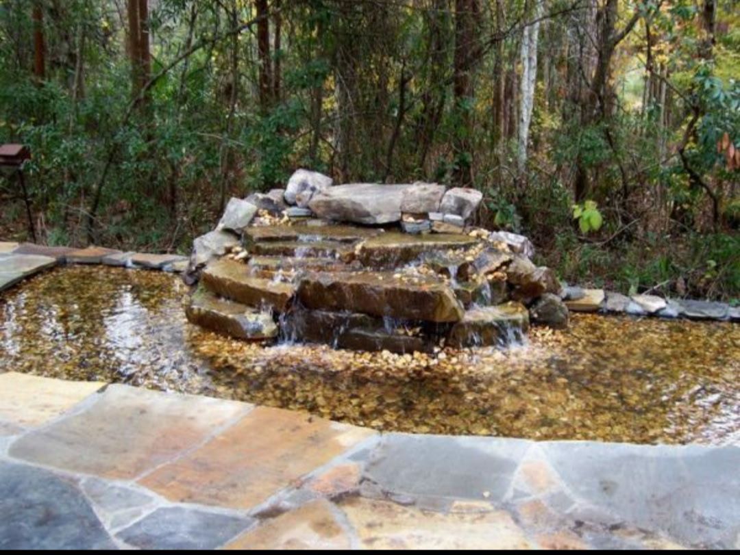 Stone waterfall cascading into a pond, surrounded by flagstone and trees.