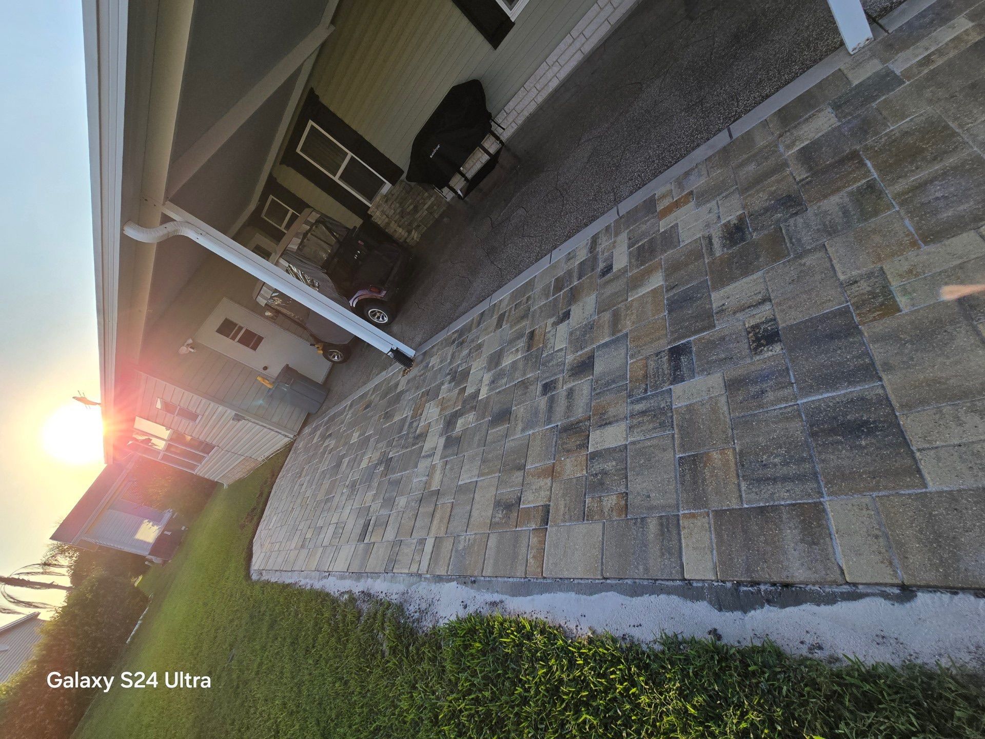 Driveway paved with rectangular stones, next to a grassy area and house with sun in the background.