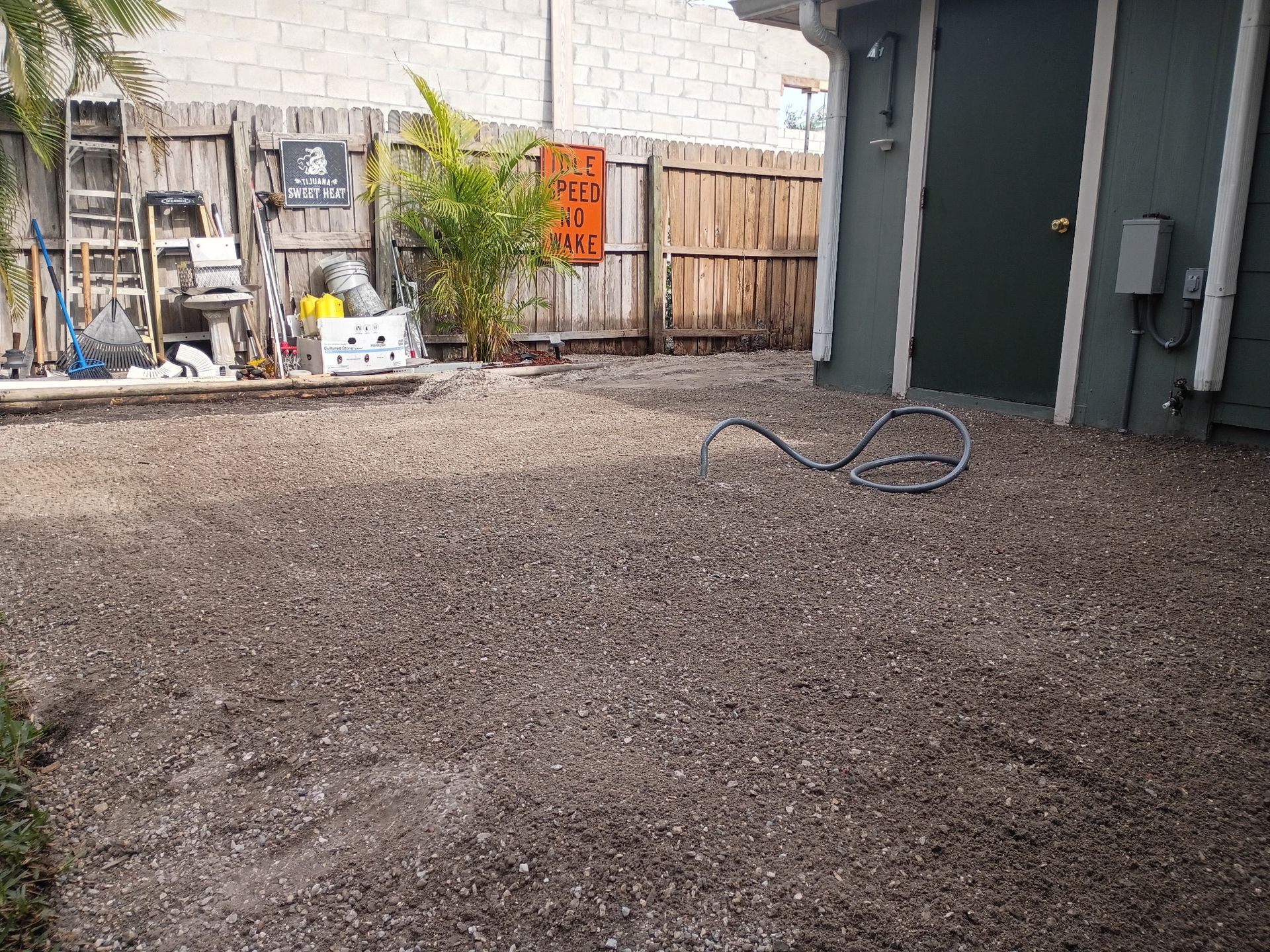 Gravel-covered backyard with a fence, palm tree, and a door. A coiled hose is on the ground.