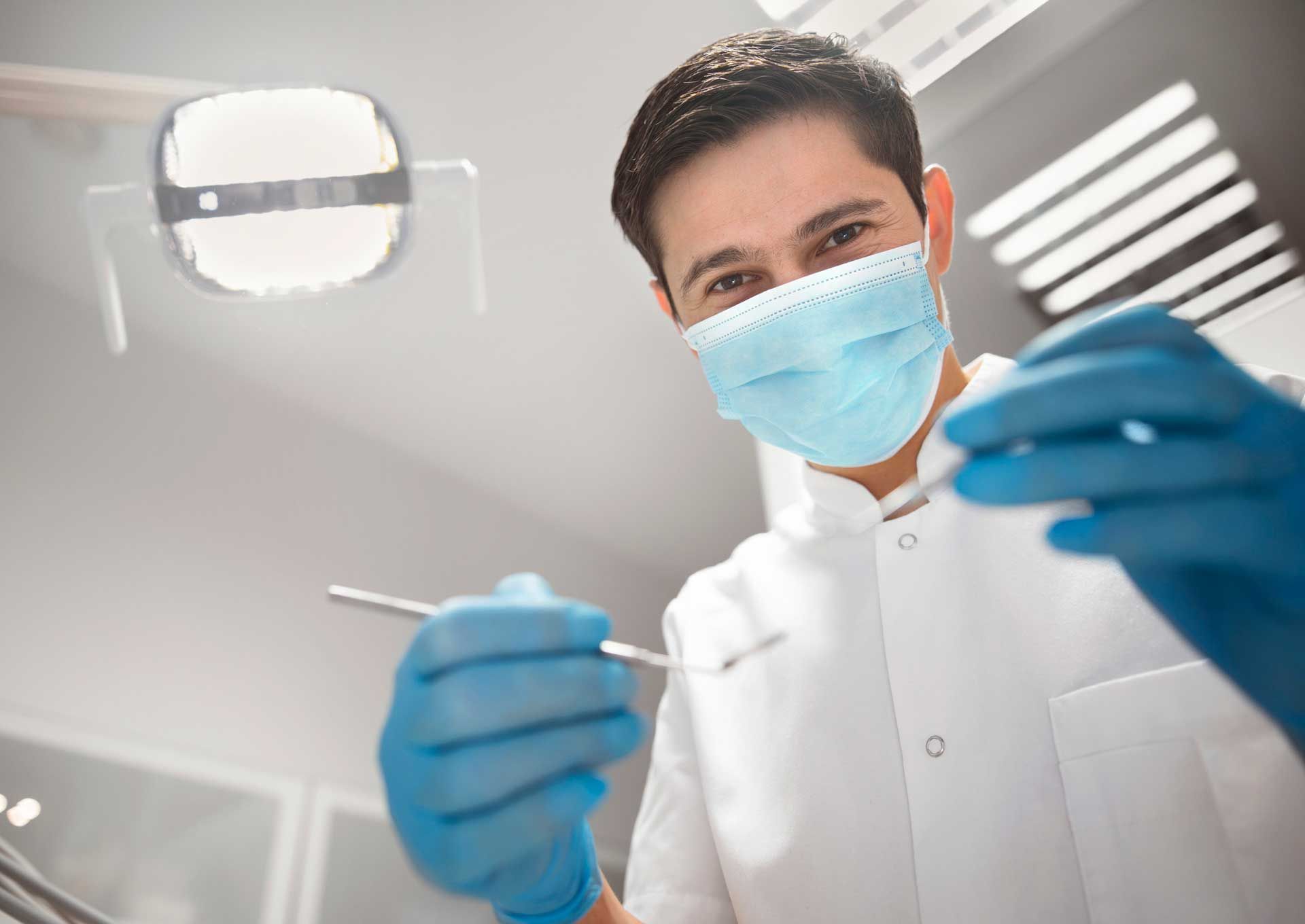Dentist wearing gloves and mask, holding dental tools in a clinic.