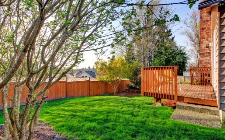 A photo of a small deck in the backyard of a house. The deck is orange/brown and sits about 3 or 4 feet off the ground. The deck is surrounded by green grass.