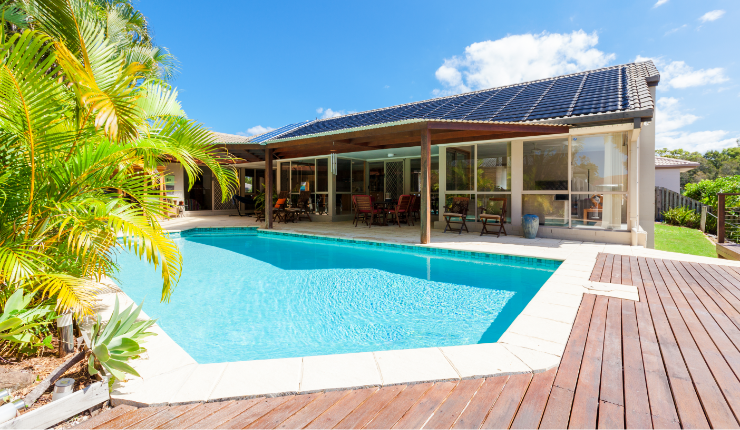 A photo of a luxurious backyard. In the backyard, there is a pergola attached to the house. Next to the pergola is a stunning in-ground pool. Surrounding the pool is a custom pool deck.