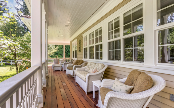 A photo of a porch in the front of a house. The house has several windows next to the porch and cushioned chairs sitting on the porch.