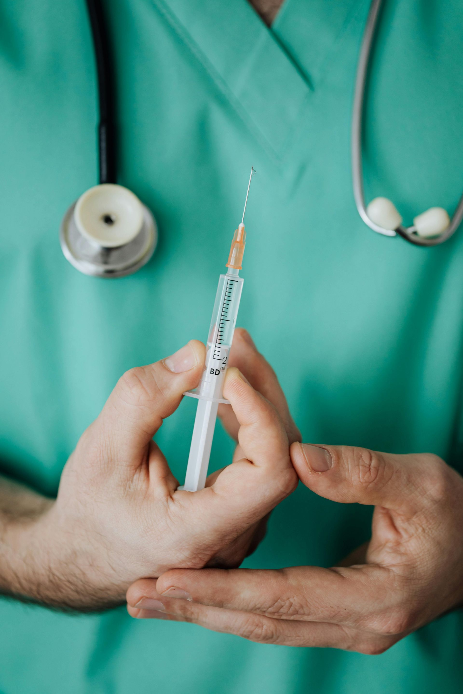 Doctor holding syringe with a needle, stethoscope around neck, wearing green scrubs.