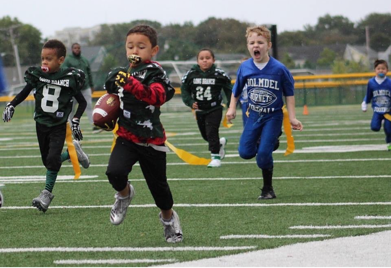 Flag football game at night, a player in red jersey runs with the ball as others pursue on a green field.