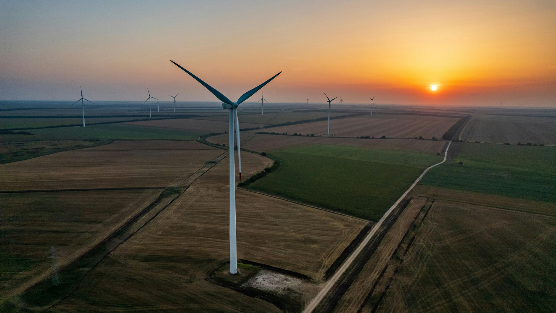 Wind turbines in a field at sunset; orange sky, green and brown fields, dirt road.