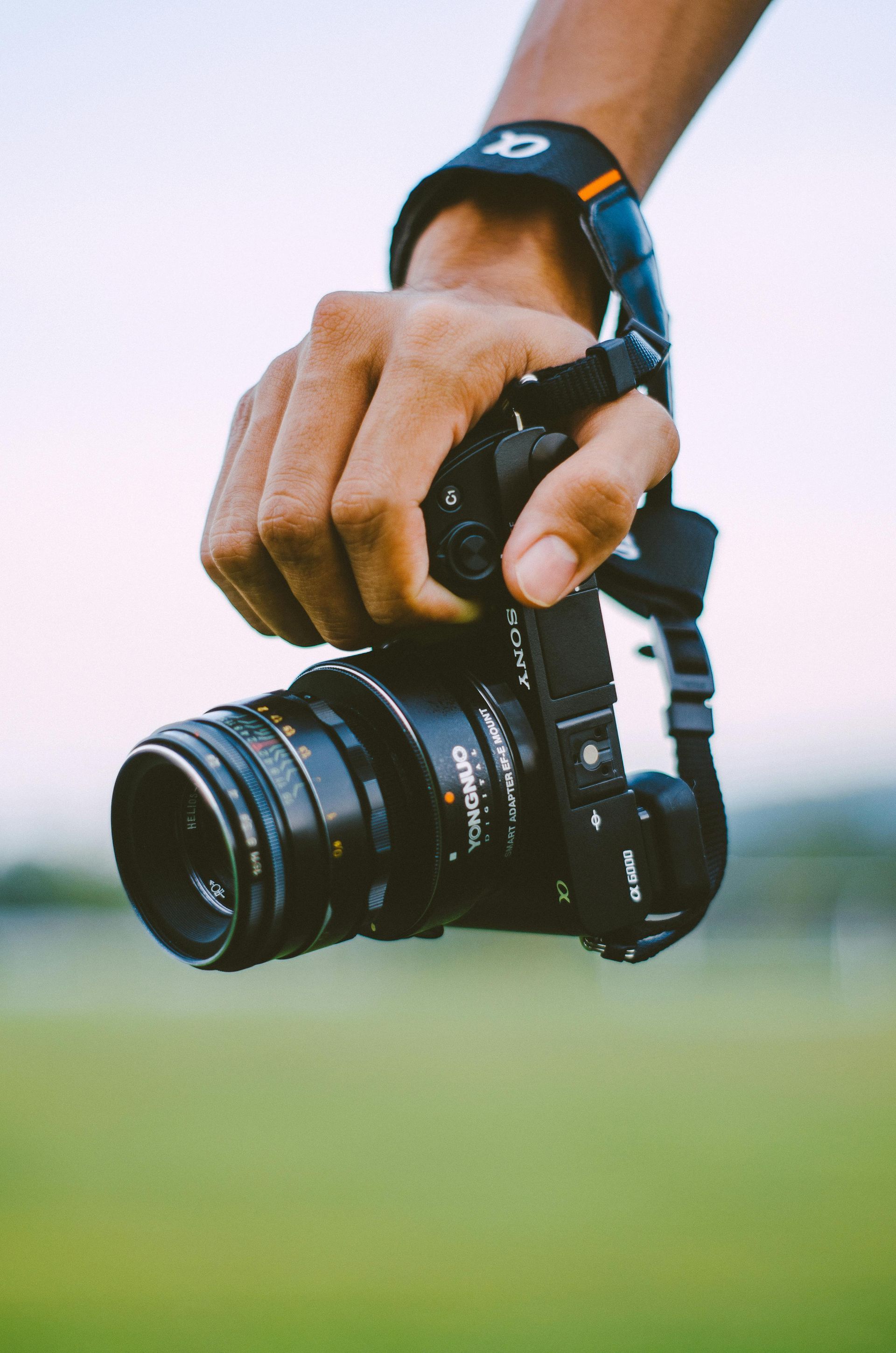 Hand holding a black camera with a lens, strap attached; outdoors, green grass in the background.