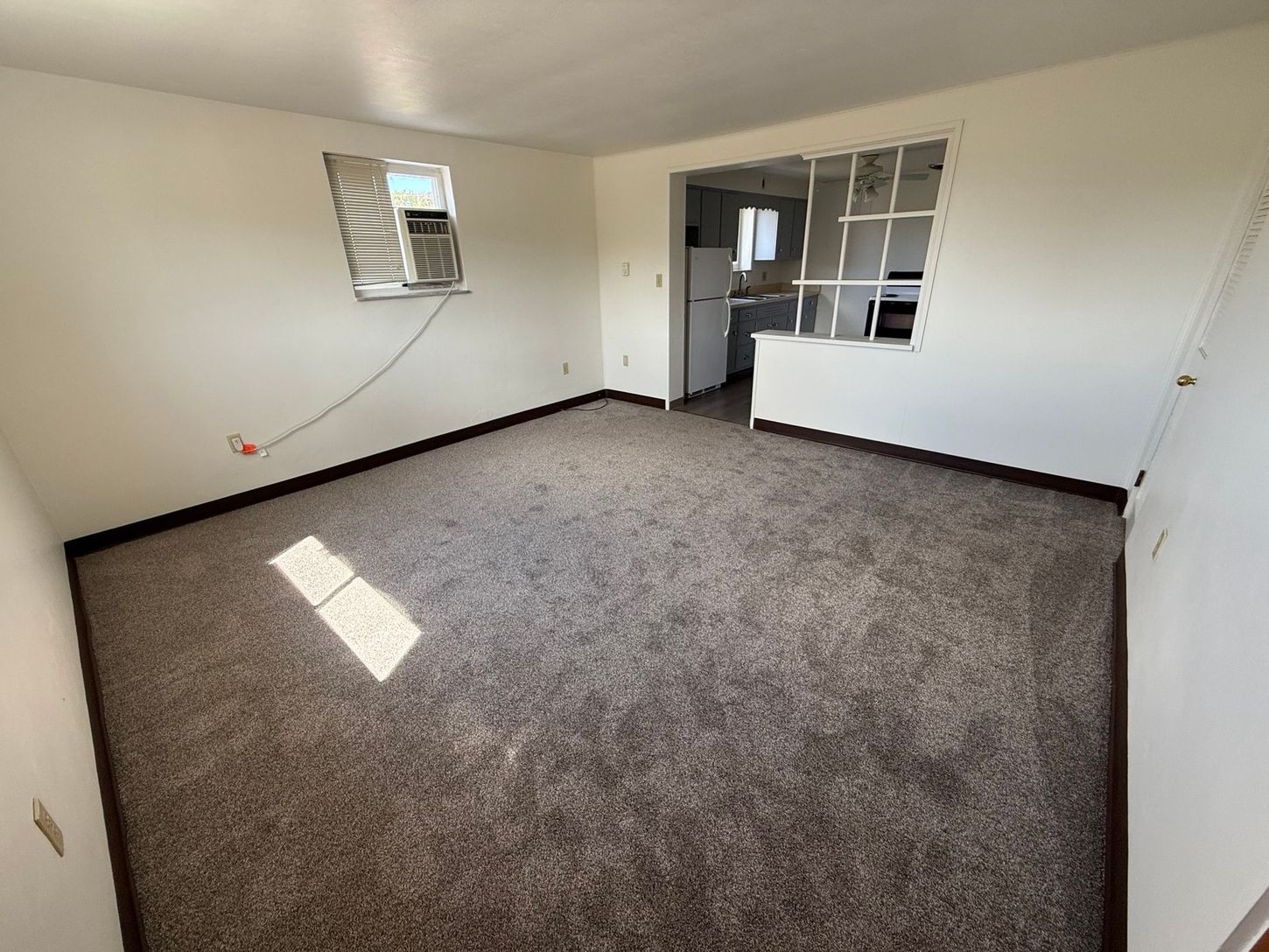 Empty room with brown carpet, white walls, and a view into a kitchen.